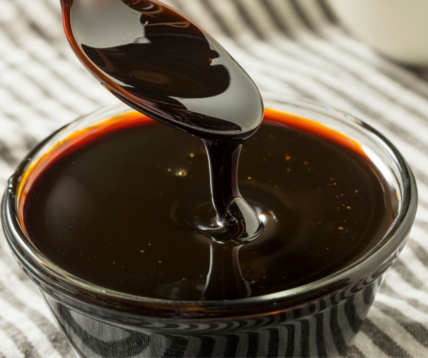 A spoonful of molasses above clear glass bowl full of dark molasses on a stripped table cloth.