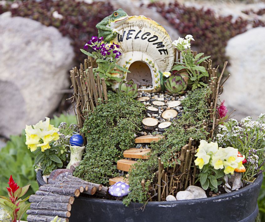 A whimsical fairy garden with a small house with the word "Welcome" on it at the top of a decorated mound.