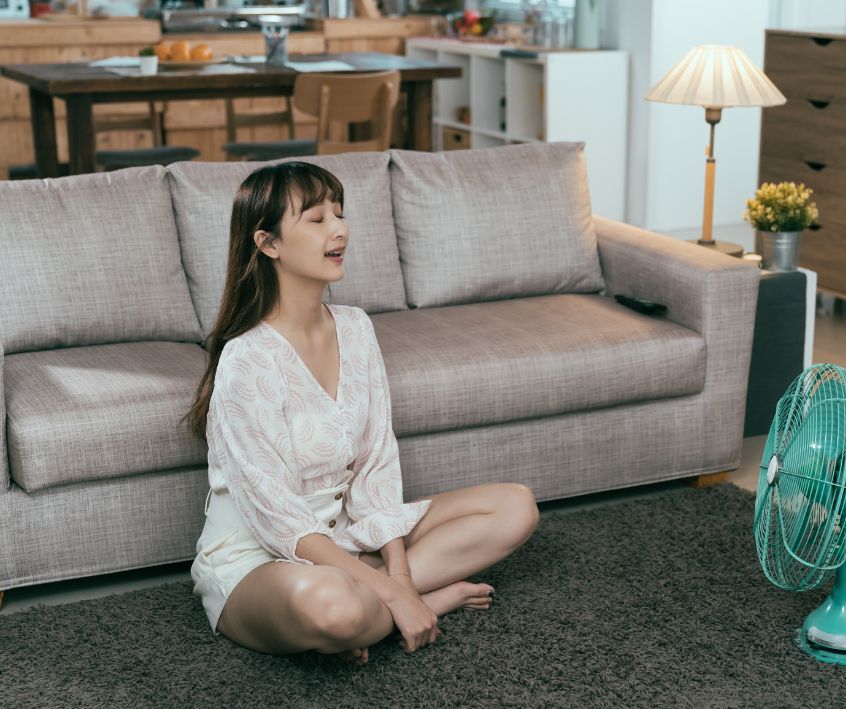 A woman in loose natural summer outfit cooling down in front of a fan.