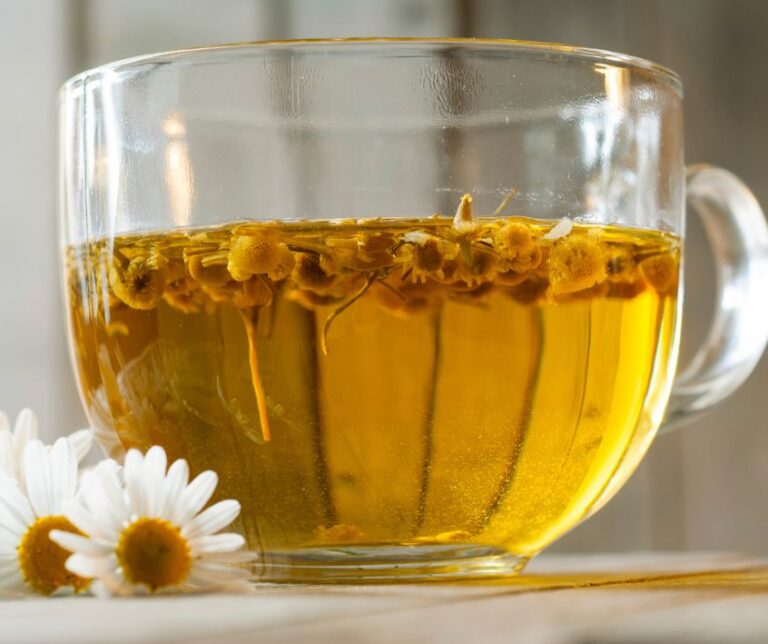 A strong brew of chamomile tea in a clear cup next to fresh chamomile flowers.