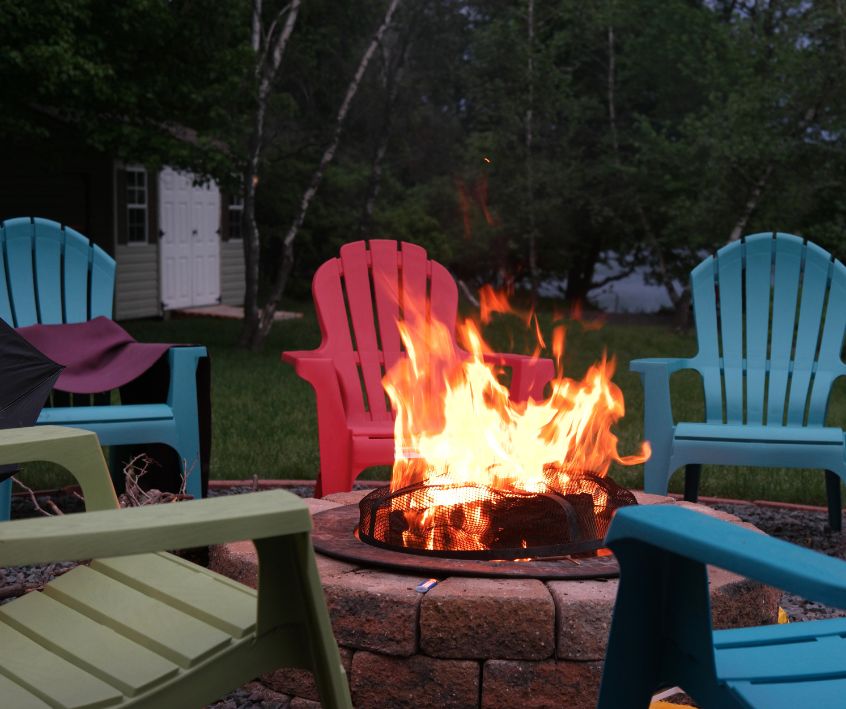 Brightly colored chair surround a stone backyard fire pit at dusk.