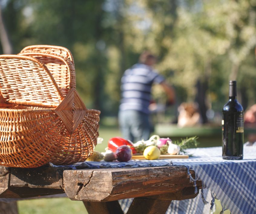 A wicker picnic basket, table cloth, and bottle of wine on a backyard picnic table.