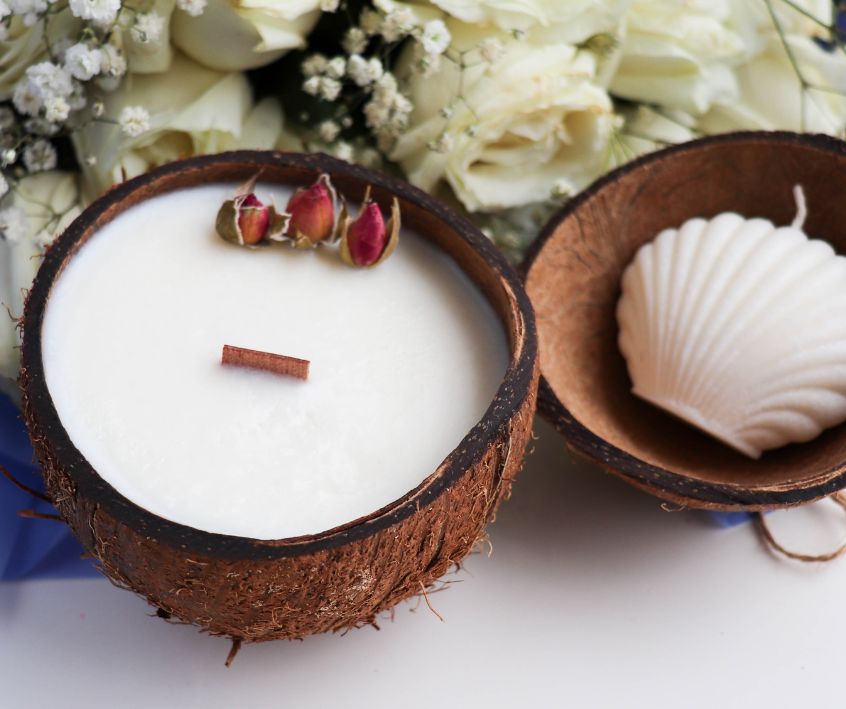 Handmade soy wax candle in coconut shell next to a white shell mold candle surrounded by white flowers.