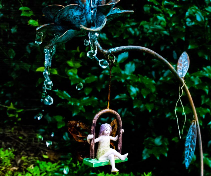 A fairy figurine on a mini flower swing a garden.