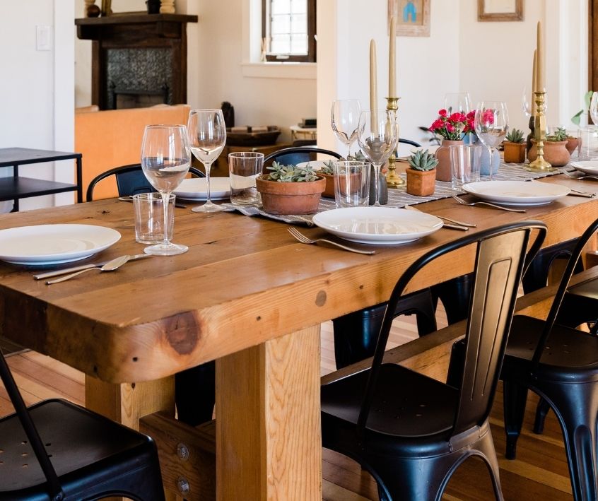 A large rustic wooden dining table with black chairs. White dishware table setting and beeswax candles in holders.