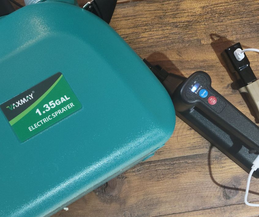 Green plastic Vaxmay 5L Electric Sprayer reservoir next to the black sprayer wand on a wooden table.