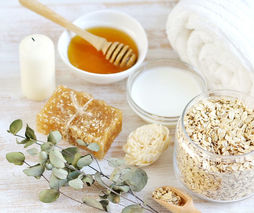 Oats, honey, milk, soap bar and candle next to a eucalyptus frond.